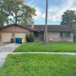Brick investment home in Jacksonville with attached two car garage and green yard with driveway showing bins outside.